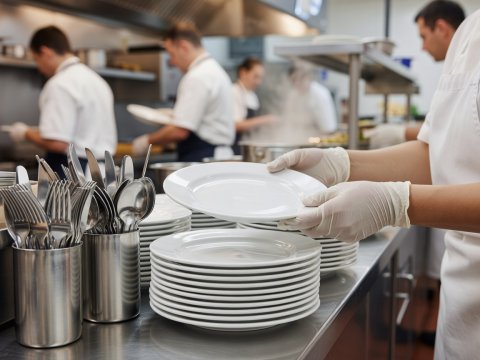 Hands stacking rolled-edge vitrified white porcelain dinner plates beside stainless flatware cylinders